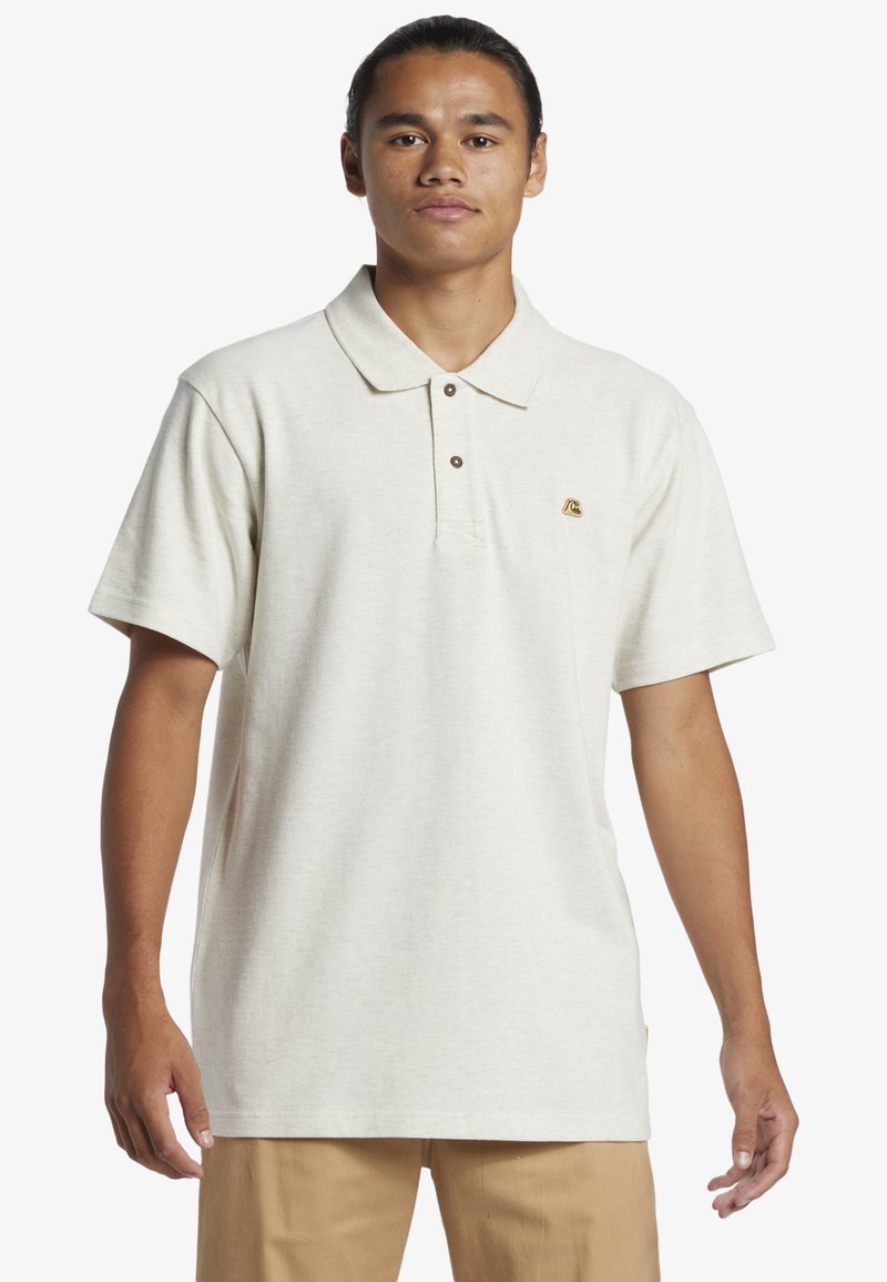Polo triko s krátkým rukávem ze světlého béžového materiálu; má límeček, zapínání na tři knoflíky a malé vyšívané logo na hrudi.