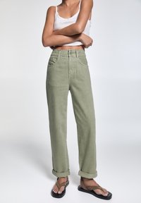 Pantalones de tiro alto, ajustados y de color verde claro, con los puños enrollados, bolsillos laterales y una tela texturizada. Combinados con sandalias planas.