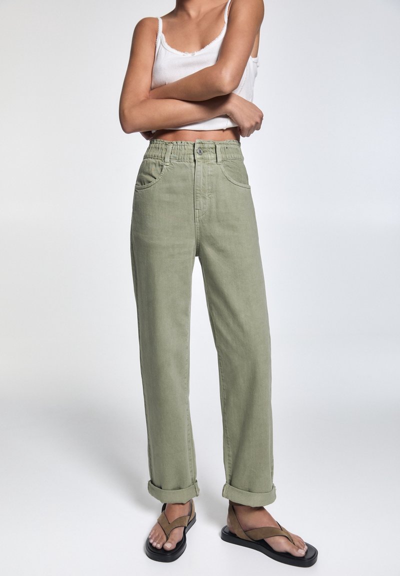 Pantalones de tiro alto, ajustados y de color verde claro, con los puños enrollados, bolsillos laterales y una tela texturizada. Combinados con sandalias planas.