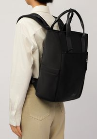 Czarny skórzany plecak z podwójnymi uchwytami u góry i regulowanymi paskami, z zamkami błyskawicznymi i kieszeniami wsuwkami, o eleganckim prostokątnym kształcie.