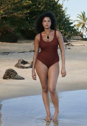 Mujer con cabello rizado natural, vistiendo un traje de baño marrón y un colgante negro, caminando descalza por una playa de arena cerca del agua y las rocas.