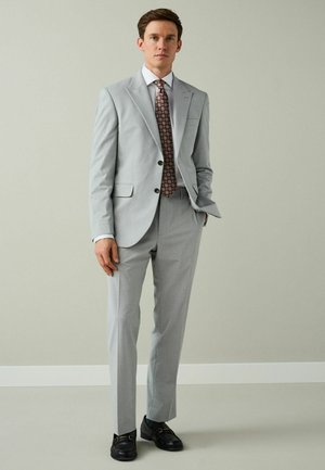 Man wearing a light gray suit with a white shirt, patterned red tie, black loafers, standing against a plain light gray background.