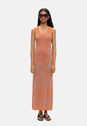 Kobieta w długiej, pomarańczowo-szarej sukience bez rękawów z wzorem, dużych okrągłych okularach przeciwsłonecznych i czarnych sandałach, stojąca na białym tle.