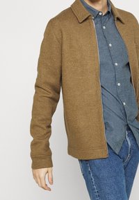 Hnědá bunda z vlněné směsi s límečkem, zapínáním na zip a žebrovanými manžetami, nošená přes světle modrou košili na knoflíky a džíny.