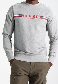 Muž v šedé mikině Tommy Hilfiger s červenými, bílými a modrými pruhy a olivových cargo kalhotách, s rukama v kapsách, hledící vpřed.