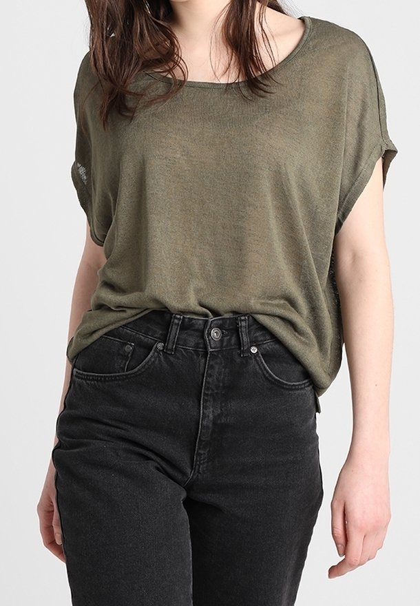 Kobieta ubrana w luźną oliwkowozieloną bluzkę z krótkim rękawem, włożoną do wysokogatunkowych czarnych dżinsów, stojąca na jednolitym tle.