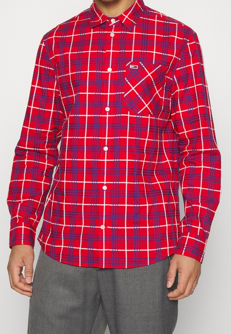 Homme portant une chemise rouge à manches longues à carreaux blancs et bleus et un pantalon gris, debout, les bras détendus.