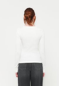 White long-sleeve ribbed top with a rounded neckline, paired with high-waisted dark jeans. The outfit features a relaxed fit and minimal detailing.