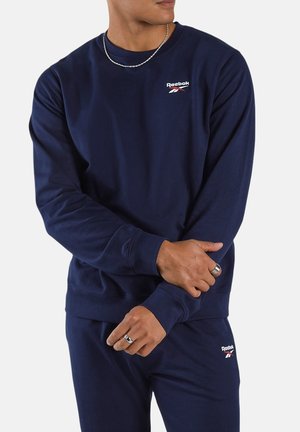 Mann trägt dunkelblauen Reebok-Pullover und passende Hose, mit silberner Kettenhalskette und Ringen, steht vor weißem Hintergrund.
