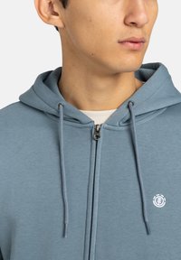 Felpa blu con zip e coulisse, realizzata in tessuto morbido, caratterizzata da un piccolo logo bianco sul petto e una texture liscia.