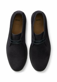 Scarpe in suede nere con un design semplice, caratterizzate da un profilo basso, punta rotonda e accenti di lacci scuri. Interno etichettato con "BOSS, MADE IN EUROPE."