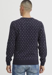 Marineblauer Strickpullover mit weißem Herzmuster, ausgestattet mit Rundhalsausschnitt und gerippten Bündchen. Der Stoff ist weich, leicht und hat eine glatte Textur.