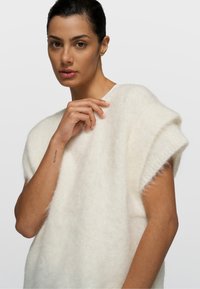 Mujer de cabello oscuro recogido, lleva un suéter blanco sin mangas y peludo, y toca ligeramente el escote contra un fondo gris liso.