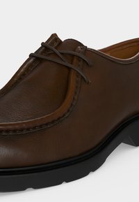 Gros plan d'une chaussure marron en cuir à lacets avec des coutures détaillées et une semelle en caoutchouc noire sur un fond clair.