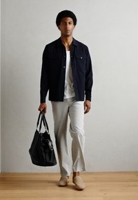 Veste boutonnière navy, t-shirt blanc, pantalon gris clair, mocassins beiges ; tient un sac fourre-tout noir ; textures lisses, design décontracté.
