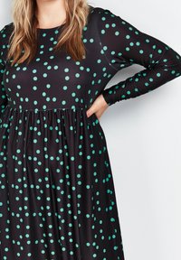 Robe noire avec un col rond et des manches longues, présentant une taille froncée et un motif de grands pois teal. Tissu extensible.