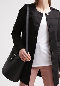 Nainen, joka on pukeutunut mustaan napitettuun takkiin valkoisen paidan päällä, pitää mustaa nahkakaulustaskua ja hänellä on päällään vaaleanpunaiset housut.