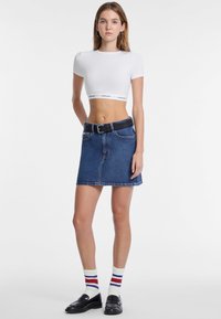 Witte cropped shirt met korte mouwen, blauwe denim rok, zwarte riem en zwarte loafers. Accessoires zijn gestreepte sokken in rood, wit en blauw.