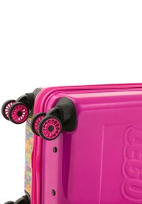 Ljust rosa bagage med en glänsande yta, försedd med små svarta hjul och ett flerfärgat mönster på sidan. Dragkedja längs toppen.