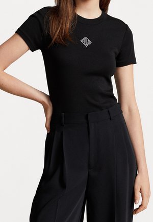 Kobieta ubrana w dopasowany czarny t-shirt z krótkim rękawem, z małym białym logo w kształcie diamentu, oraz czarne, plisowane spodnie z wysokim stanem.