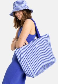 Blauwe en witte gestreepte tote bag met een gerimpelde bovenkant, gedragen door een persoon in een blauwe mouwloze jurk en bijpassende hoed.