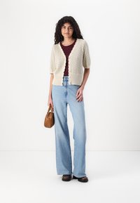 Short-sleeved cream cardigan with textured pattern, scalloped edges, and five buttons, paired with high-waisted blue wide-leg jeans and a brown bag.