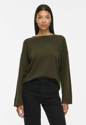 Femme aux longs cheveux noirs portant un pull vert foncé ample et un pantalon noir, debout devant un fond blanc uni.