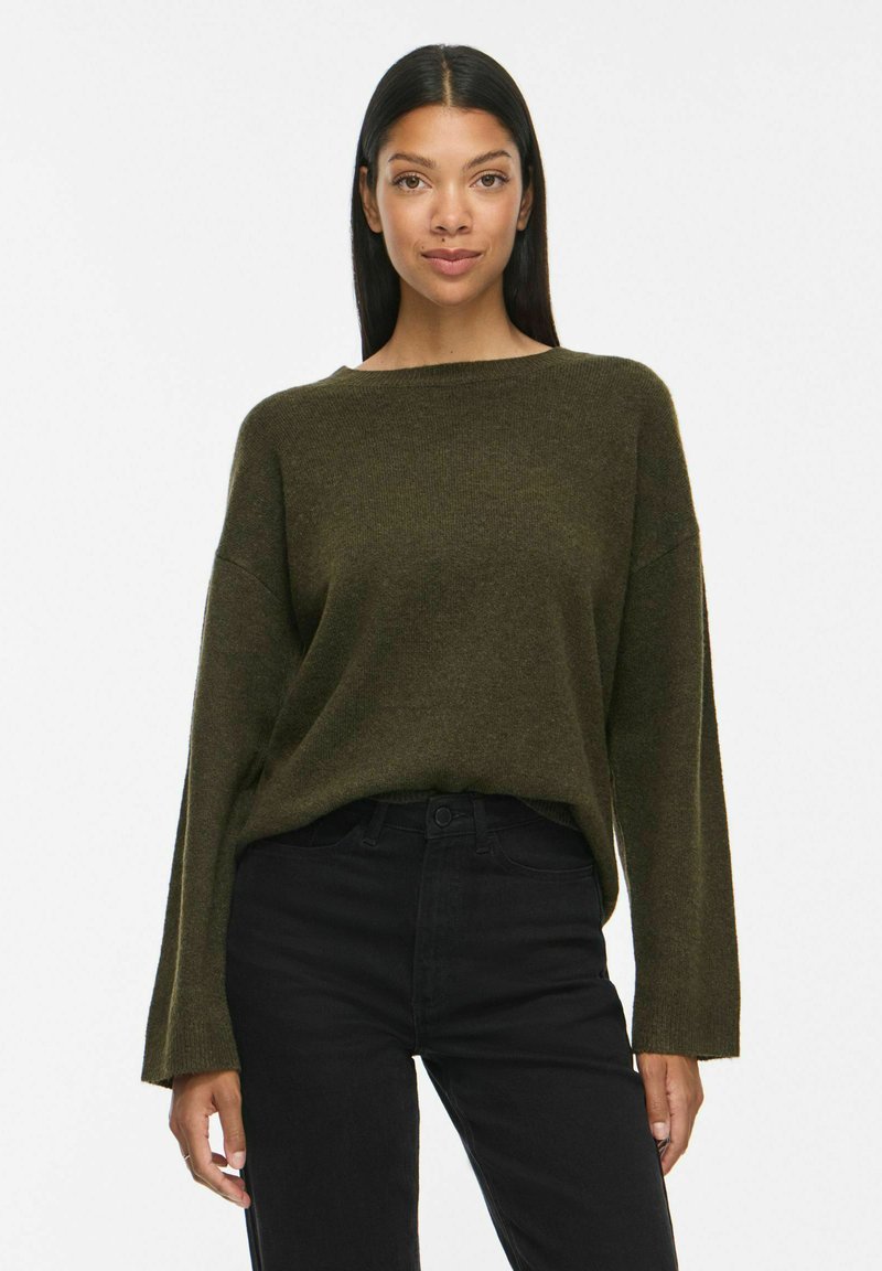 Femme aux longs cheveux noirs portant un pull vert foncé ample et un pantalon noir, debout devant un fond blanc uni.