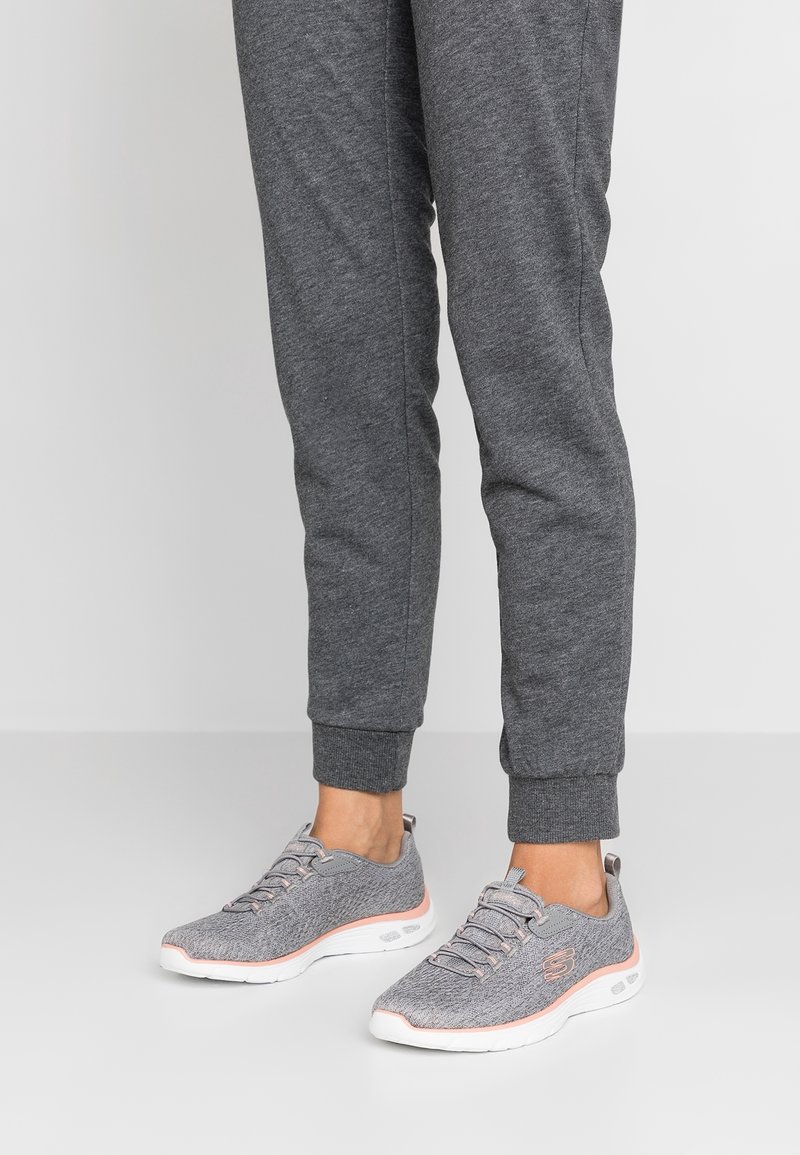 Grå idrottsskor med en texturerad ovandel, vit sula och rosa detaljer. Bärs med grå joggingbyxor som har en åtsittande mudd vid ankeln.