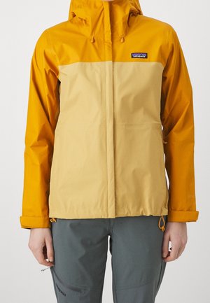 Osoba w dwukolorowej, musztardowo-żółtej i beżowej kurtce z kapturem Patagonia oraz ciemnoszarych spodniach outdoor, stojąca na prostym tle.