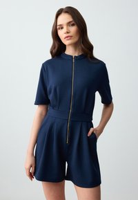 Navyblauw kort jumpsuit van een zachte stof, met een kraag, korte mouwen en een gouden rits. Inclusief zijzakken en een getailleerde taille.