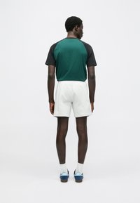 Camisa de mangas curtas com blocos de cor verde e preto, combinada com calções brancos e sapatos azuis. Vista de costas, destacando o design simples e o ajuste.