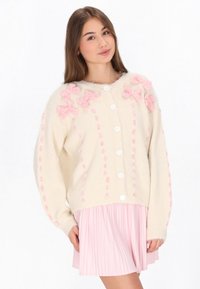 Crème cardigan met roze getextureerde strikken, grote witte knopen en verticale gebreide patronen. Gecombineerd met een lichtroze geplisseerde rok.
