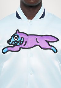 Lichtblauwe satijnen jas met contrasterende marineblauwe geribde kraag, met een grote paarse en blauwe cartoonkat geborduurd op de voorkant.