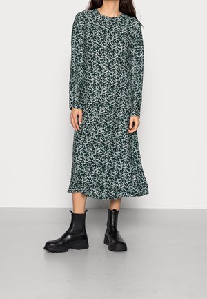 Femme portant une robe midi à manches longues, fleurie vert foncé, associée à des bottines noires épaisses, debout sur un sol gris.