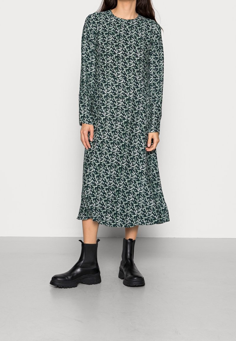 Femme portant une robe midi à manches longues, fleurie vert foncé, associée à des bottines noires épaisses, debout sur un sol gris.