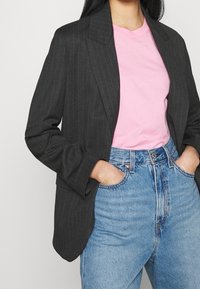 Schwarzer Blazer mit Nadelstreifen und taillierter Passform, getragen über einem rosa T-Shirt, kombiniert mit hochgeschnittenen blauen Jeans. Bemerkenswerte Details sind die strukturierten Schultern.