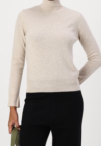 Beige polotröja i mjukt stickat tyg, med långa ärmar och ribbade muddar och fåll. Matcherad med svarta byxor, hållna ihop med ett grönt band.