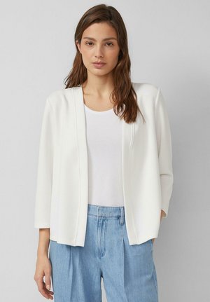 Junge Frau mit langen braunen Haaren, die eine weiße offene Jacke, ein weißes Oberteil und hellblaue, hoch taillierte Jeans trägt, vor einem schlichten grauen Hintergrund steht.