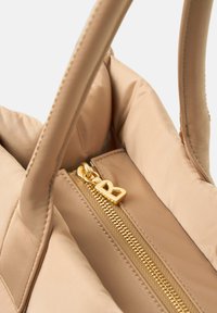 Beige quiltet håndtaske indeni med en gylden lynlås og en lynlåsgreb formet som et 'R', udstyret med to håndtag og en glat stoftekstur.