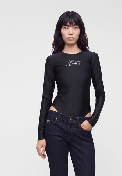 Femme aux longs cheveux noirs portant un body noir à manches longues et un jean bleu foncé, debout devant un fond clair uni.