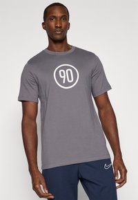 T-shirt grigio a maniche corte realizzato in cotone, con una grafica circolare bianca che presenta il numero "90" al centro. Vestibilità casual e design semplice.