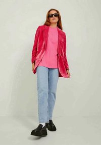 Fløyelsrød blazer, rosa t-skjorte, lyseblå jeans og sorte platåsko. Modellen har på seg sorte solbriller og står mot en nøytral bakgrunn.