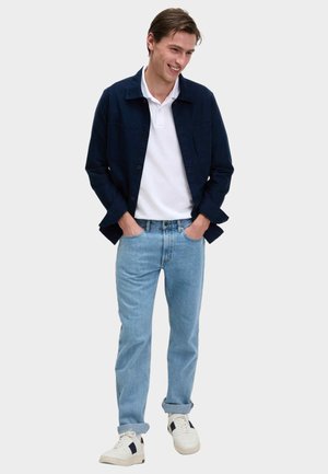 Junger Mann lächelt, trägt eine marineblaue Jacke über einem weißen Poloshirt, hellblaue Jeans mit hochgekrempelten Bündchen und weiße Turnschuhe mit schwarzen Akzenten.