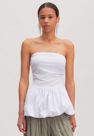 Vrouw draagt een witte strapless peplum top en een lange olijfgroene geplooide rok, met lange hangende pareloorbellen, staand tegen een effen achtergrond.