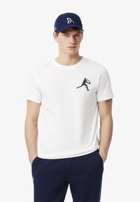 Camiseta blanca de manga corta con un gráfico oscuro de un jugador en movimiento en el lado izquierdo del pecho, combinada con un pantalón azul marino y una gorra azul marino.