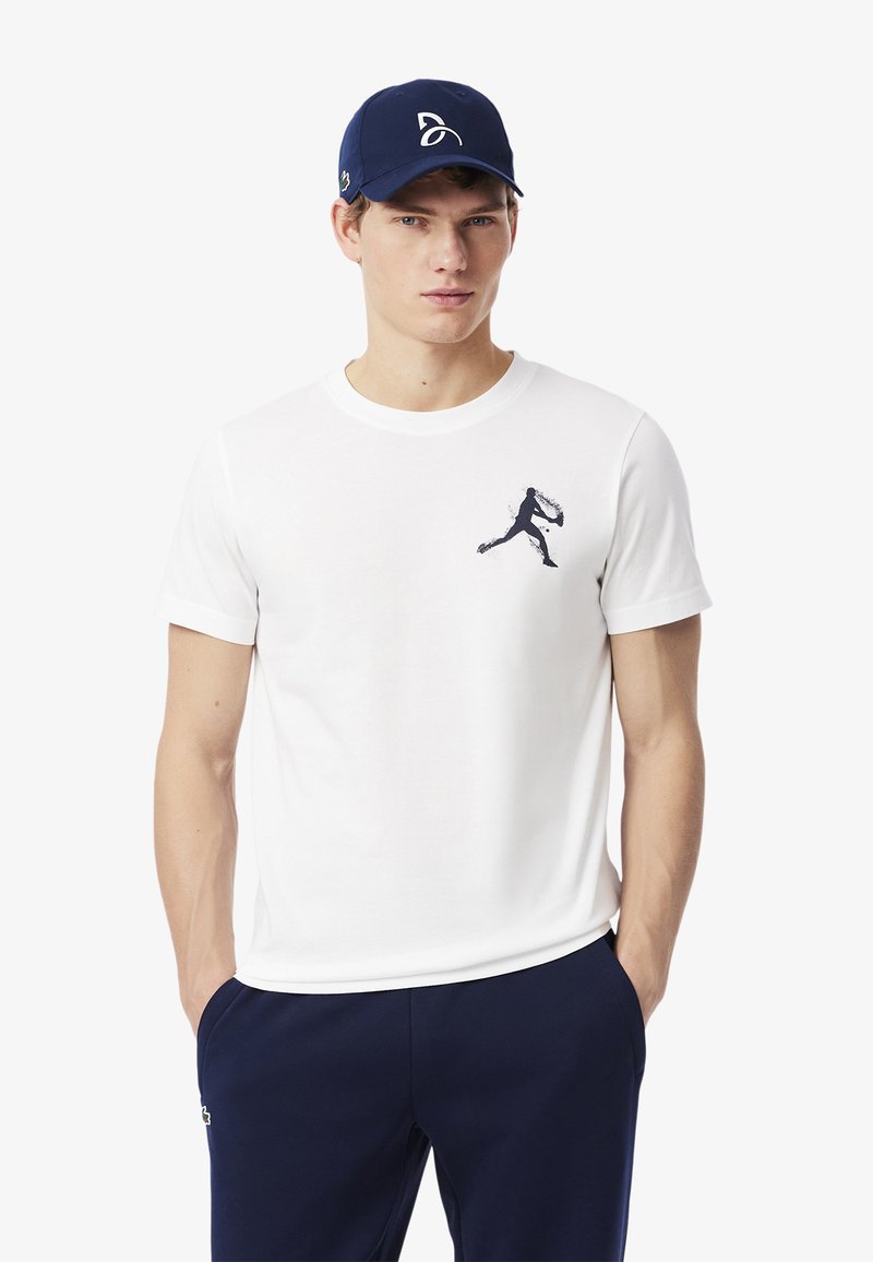 Camiseta blanca de manga corta con un gráfico oscuro de un jugador en movimiento en el lado izquierdo del pecho, combinada con un pantalón azul marino y una gorra azul marino.