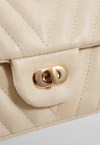 Beige nahkakäsilaukku, jossa on päällänsä tikattu kuvio, varustettuna kullansävyisellä metallilla varustetulla painonappisuljenta ja huomaamattomilla tikkaus yksityiskohdilla.