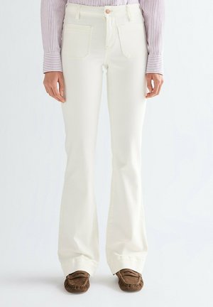 Person iført cremefarvede bukser med høj talje og vide ben, brune mokkasinsko og en langærmet, stribet skjorte med hænder afslappet ned langs siderne.