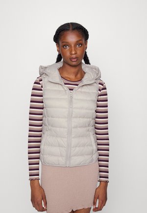 Gilet bouffant gris clair avec capuche, fermeture éclair à l'avant, et texture matelassée. Porté par-dessus un haut à manches longues rayé avec des rayures violettes et blanches.